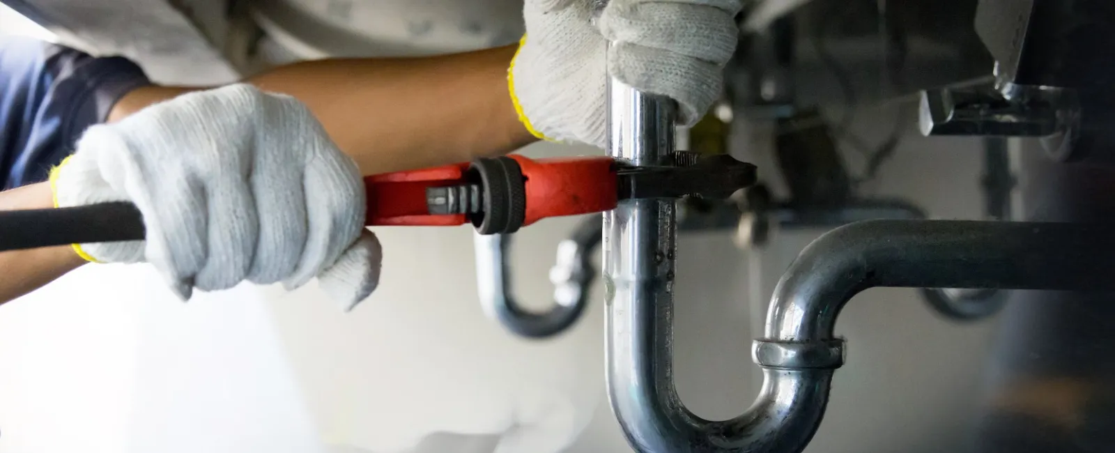 Plumber repairing pipes under a sink with a wrench and gloves.