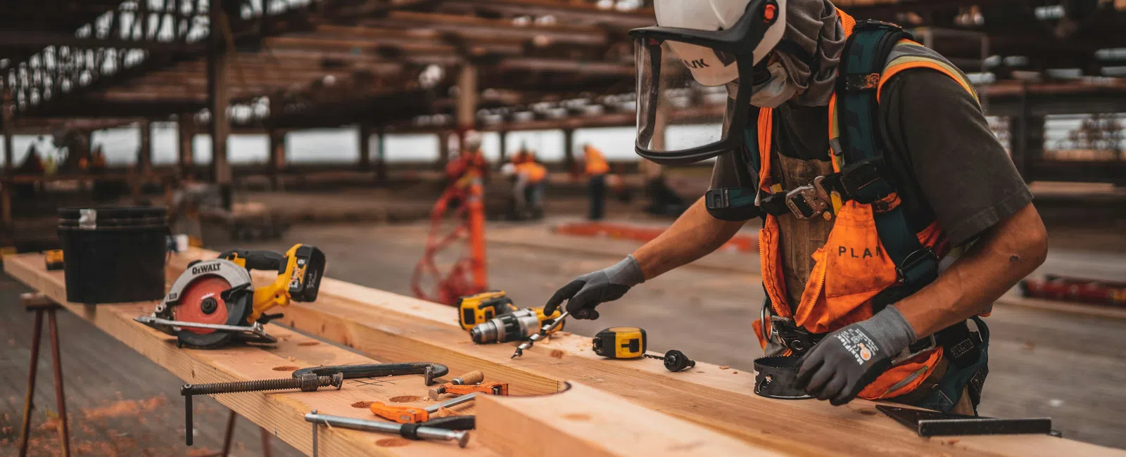 Construction worker wearing safety gear carefully measuring wood on site with tools and power saw in background.