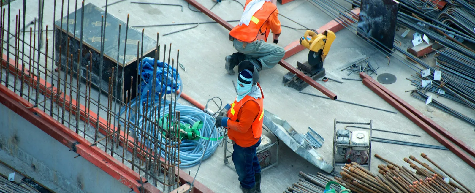 Construction workers in orange vests and helmets working on steel reinforcement at a building site.