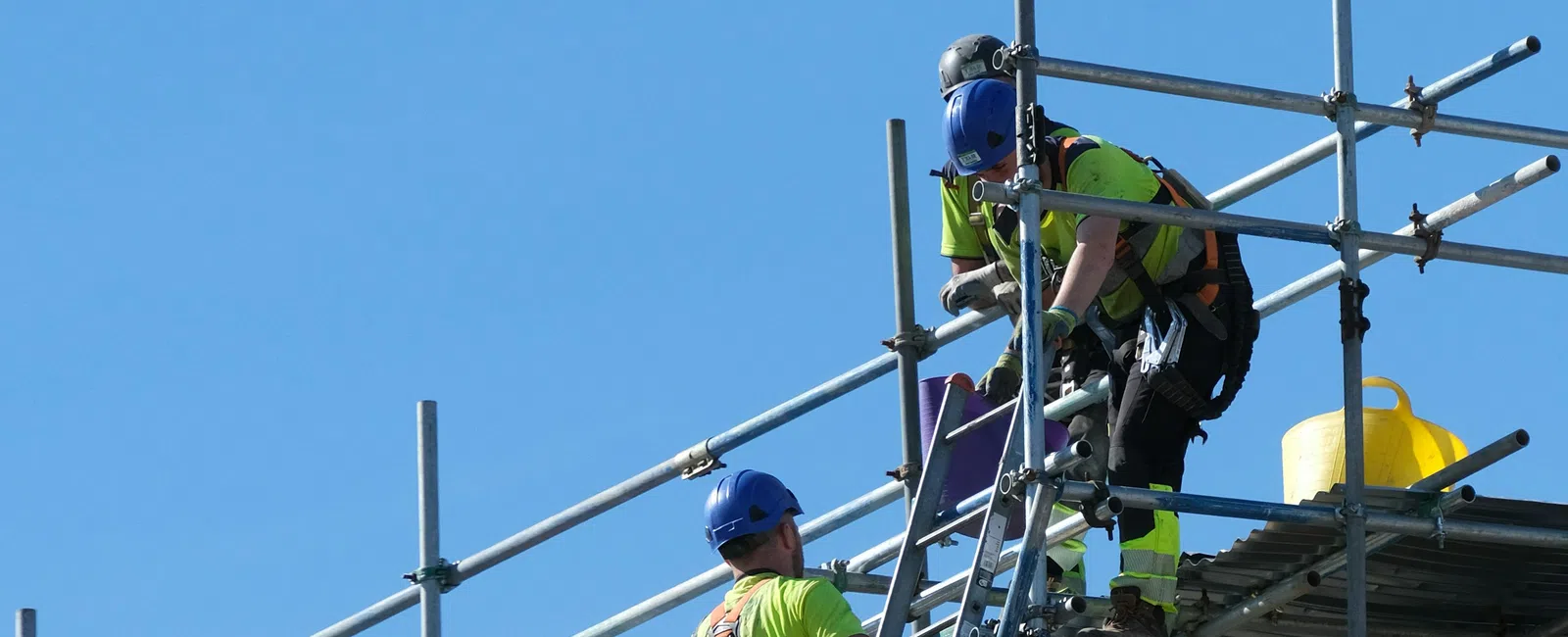 Construction workers wearing safety gear assembling scaffolding against a clear blue sky.