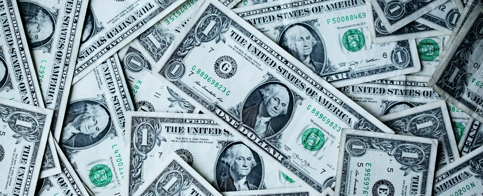 Pile of US one dollar bills overlapping showing George Washington portraits and green seals.