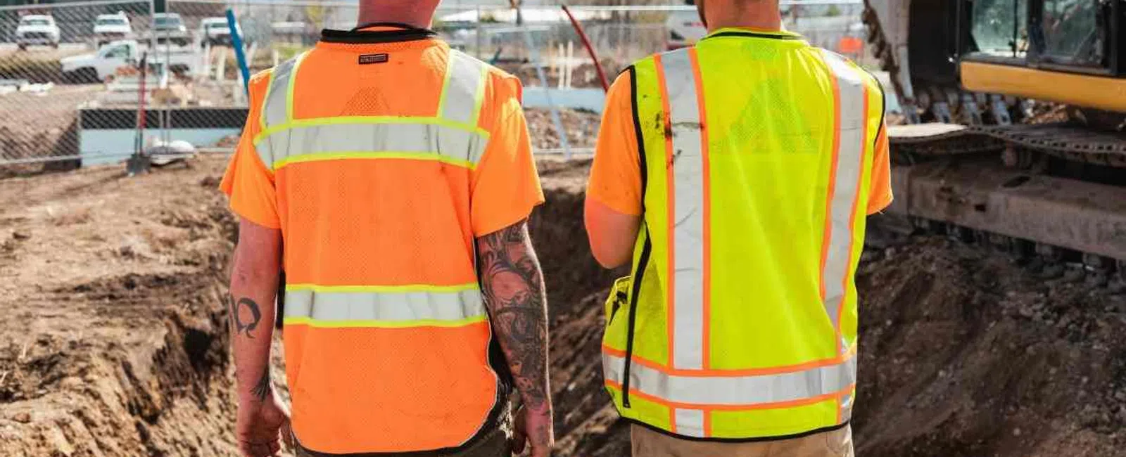 Two construction workers in safety vests and helmets standing at an excavation site near heavy machinery.