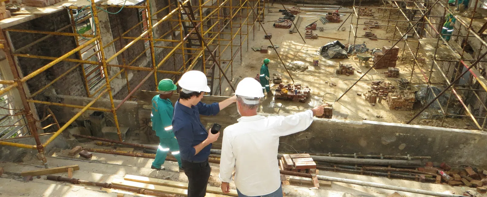 Two construction supervisors in hard hats discussing site plans on a building construction site with scaffolding.