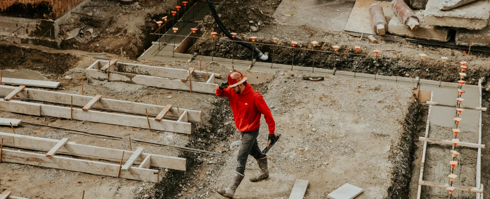 Construction worker in a red jacket and hard hat walking across a foundation site with gravel and wooden frames.