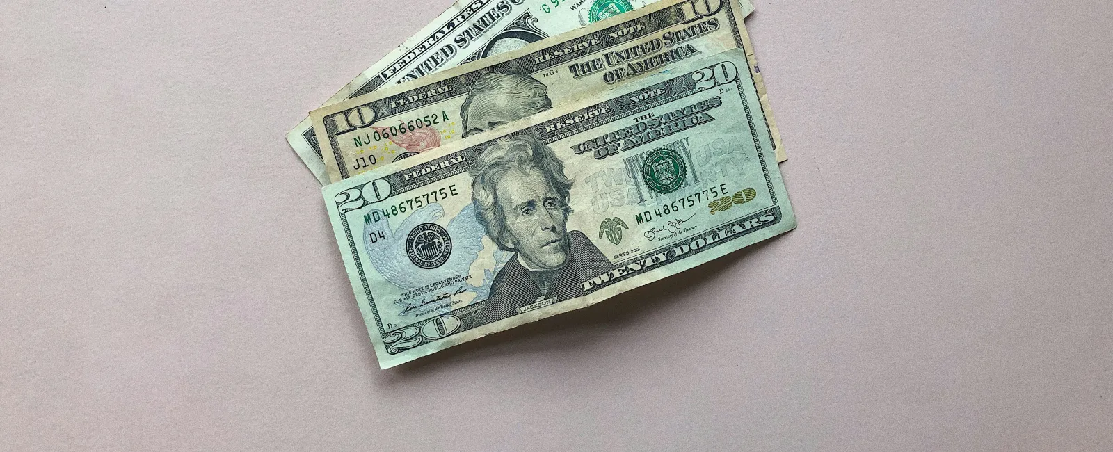 Two US dollar bills, one twenty-dollar and one one-dollar, on a plain light pink surface.