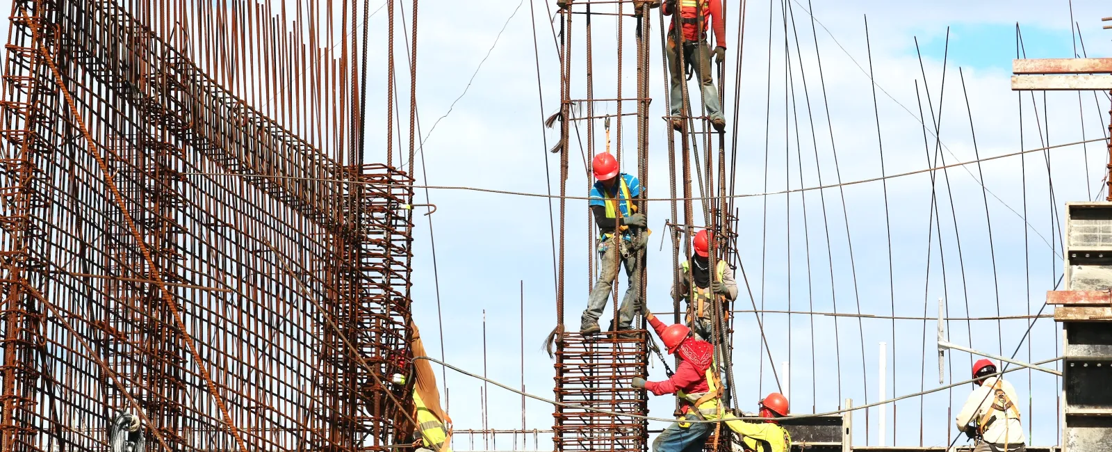 Construction workers wearing safety gear working on steel rebar framework at a building site under clear sky.