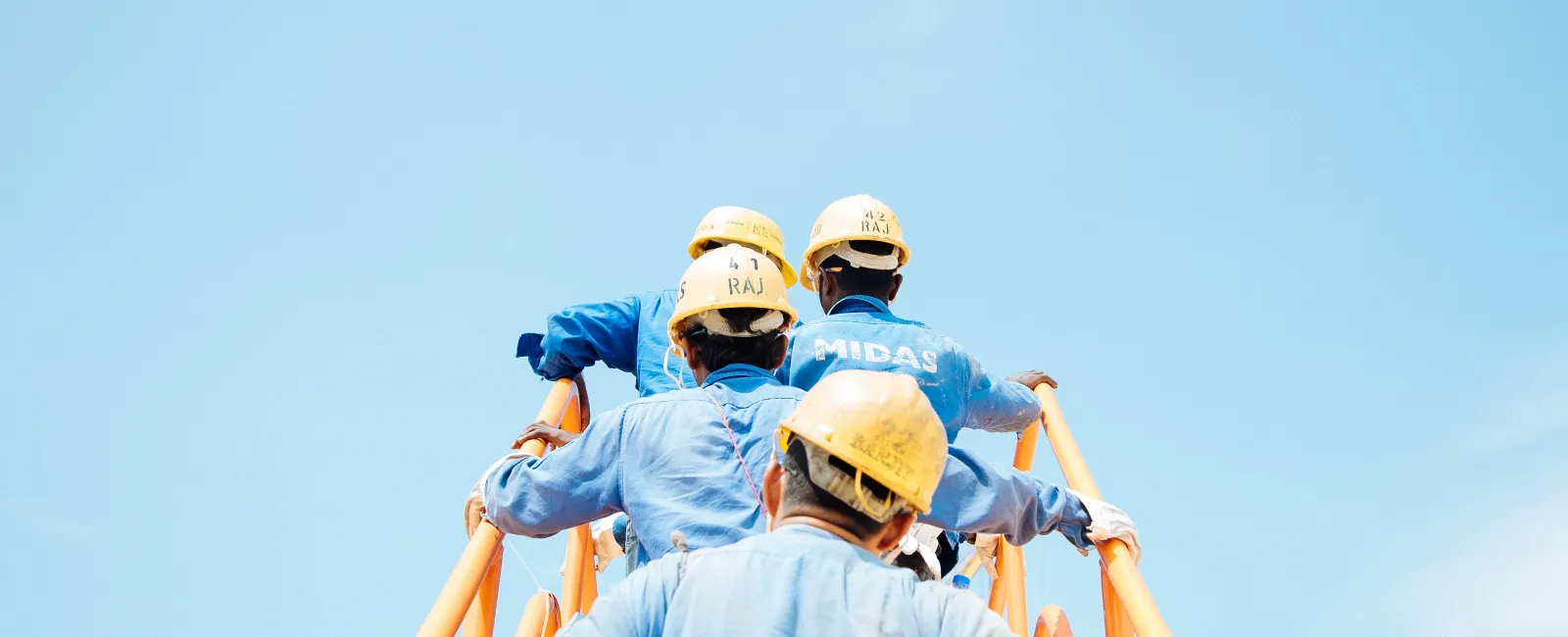 Construction workers wearing yellow helmets and blue uniforms climbing orange metal stairs under a clear blue sky