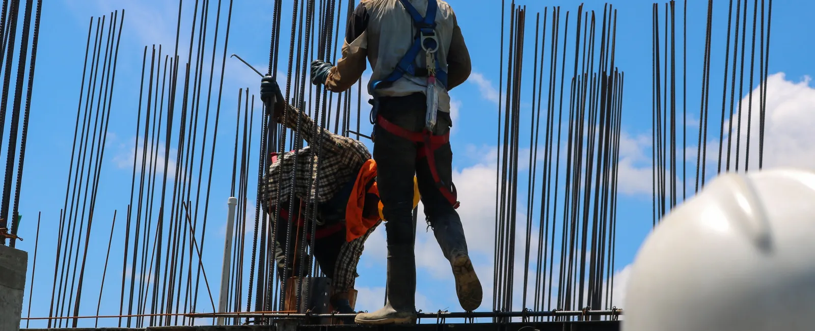 Construction workers wearing safety gear working amidst steel rebar framework under a blue sky.