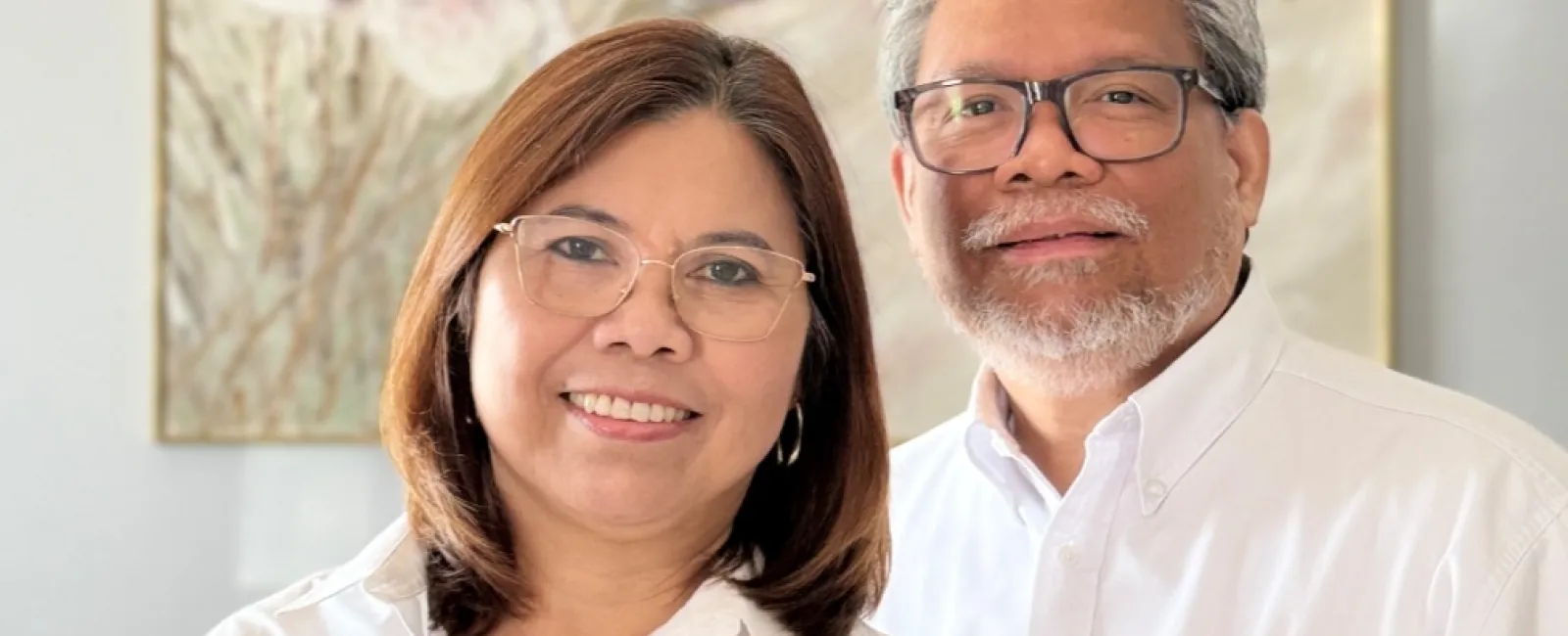 Smiling man and woman wearing white trustbridge shirts stand in front of floral wall art indoors.