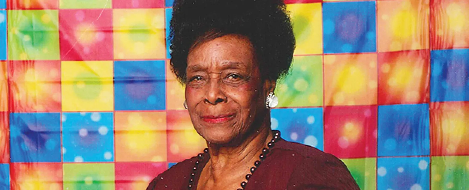 Dr. Bernice Catherine Harper in maroon outfit posed with hands raised against colorful checkered backdrop.