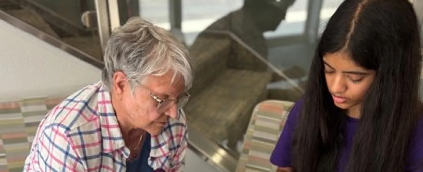 Older woman teaching young girl to crochet while seated on striped chairs with yarn containers on the floor.