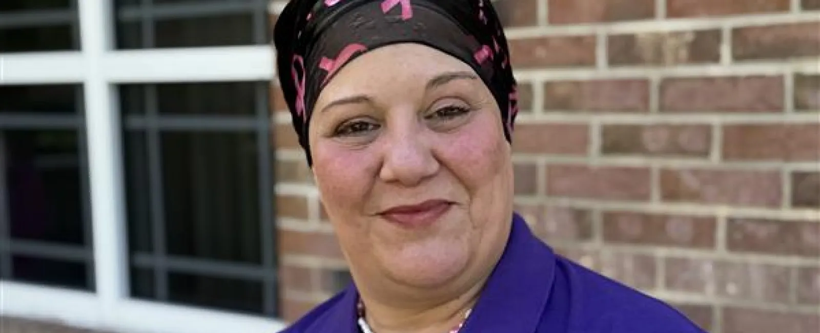 Smiling woman in a purple Hospice of Marion County polo and pink ribbon headscarf stands outside a brick building.