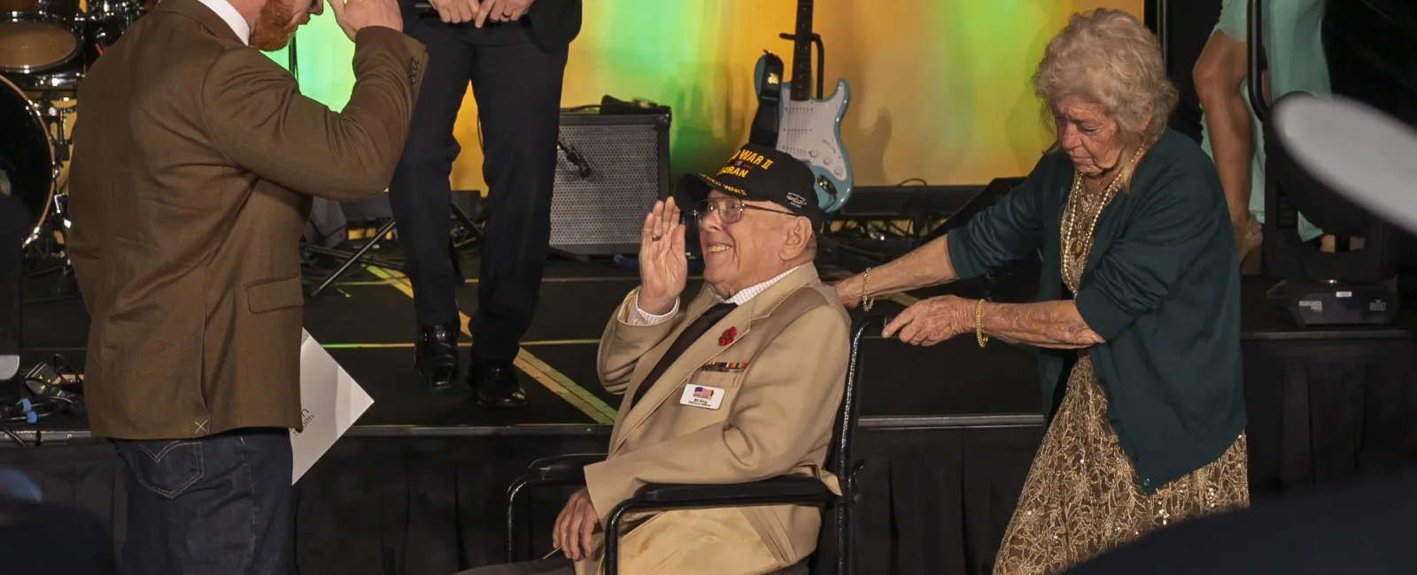 Veteran in wheelchair salutes man standing nearby as elderly woman pushes wheelchair at event stage with musical instruments.