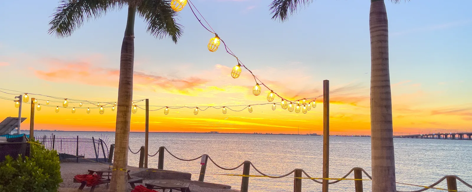 Beachside picnic area with palm trees, string lights, and sunset over calm water with a distant bridge.
