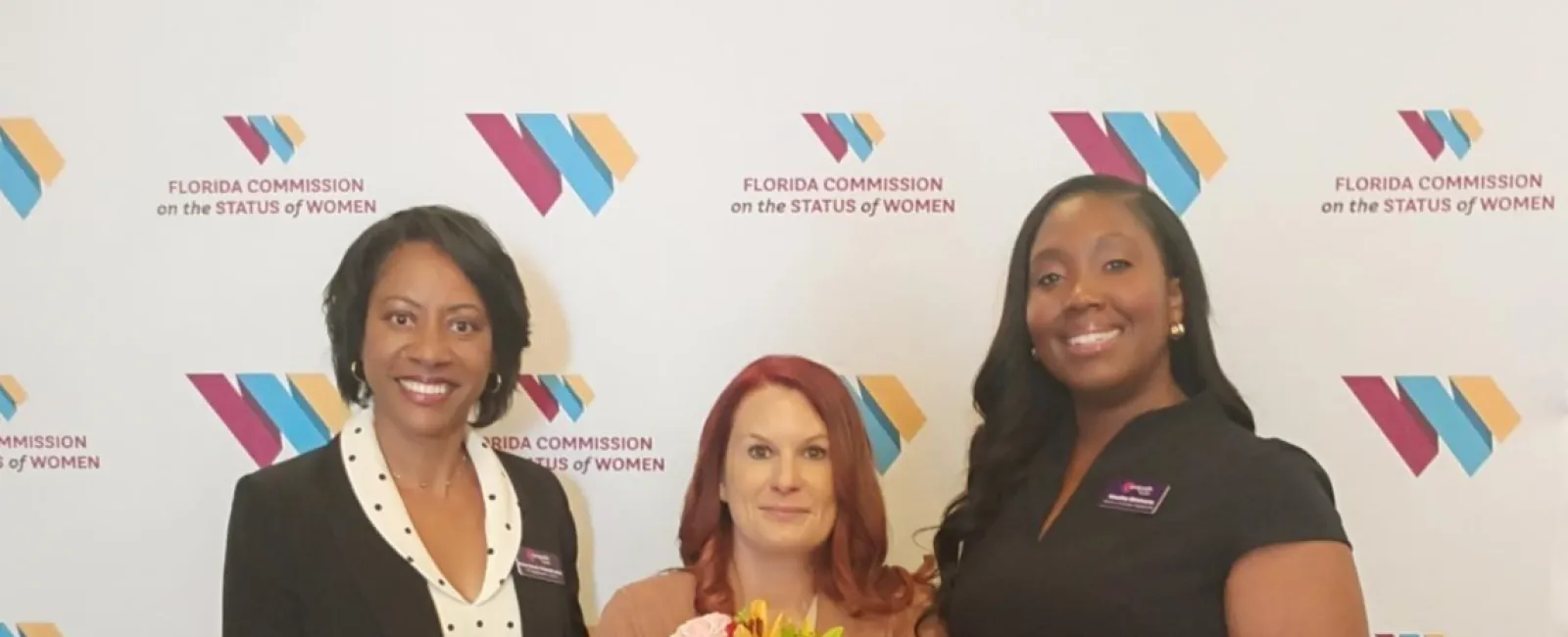 Three women standing in front of Florida Commission on the Status of Women backdrop, one holding a flower bouquet.