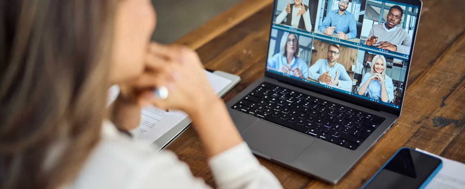 Woman in white shirt attending a virtual meeting with six diverse colleagues on a laptop at a wooden table.