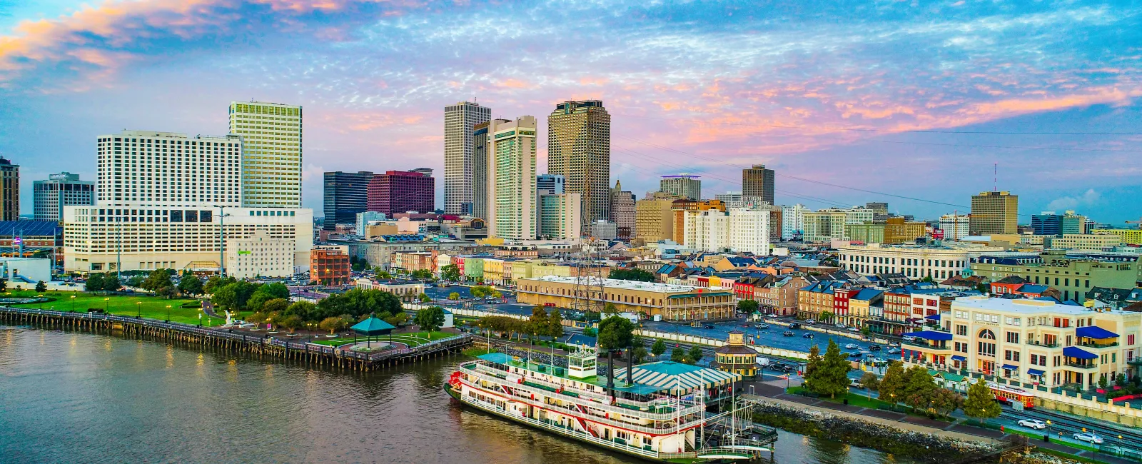 City skyline with high-rise buildings, riverfront park, and a paddlewheel riverboat at sunset under a colorful sky.