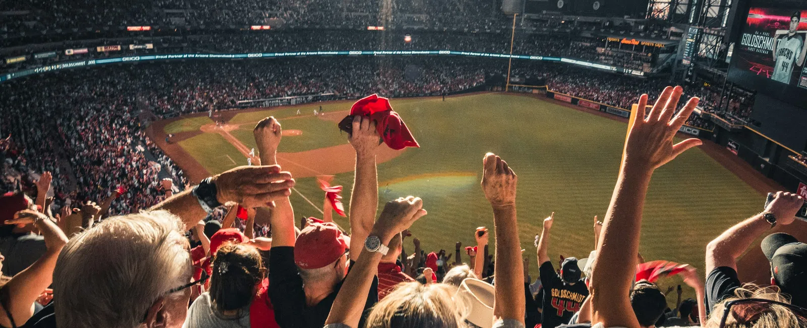 Crowd of fans cheering with raised hands inside a sunlit indoor baseball stadium during a game.