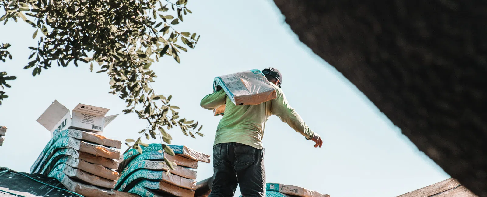 Worker carrying roofing material on a wooden rooftop with stacked shingles and tree branches in the background