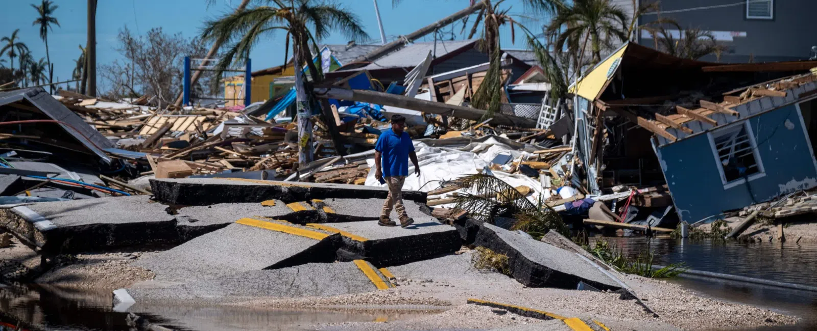 Man walks on collapsed road amid destroyed homes and debris after hurricane damage under clear blue sky.