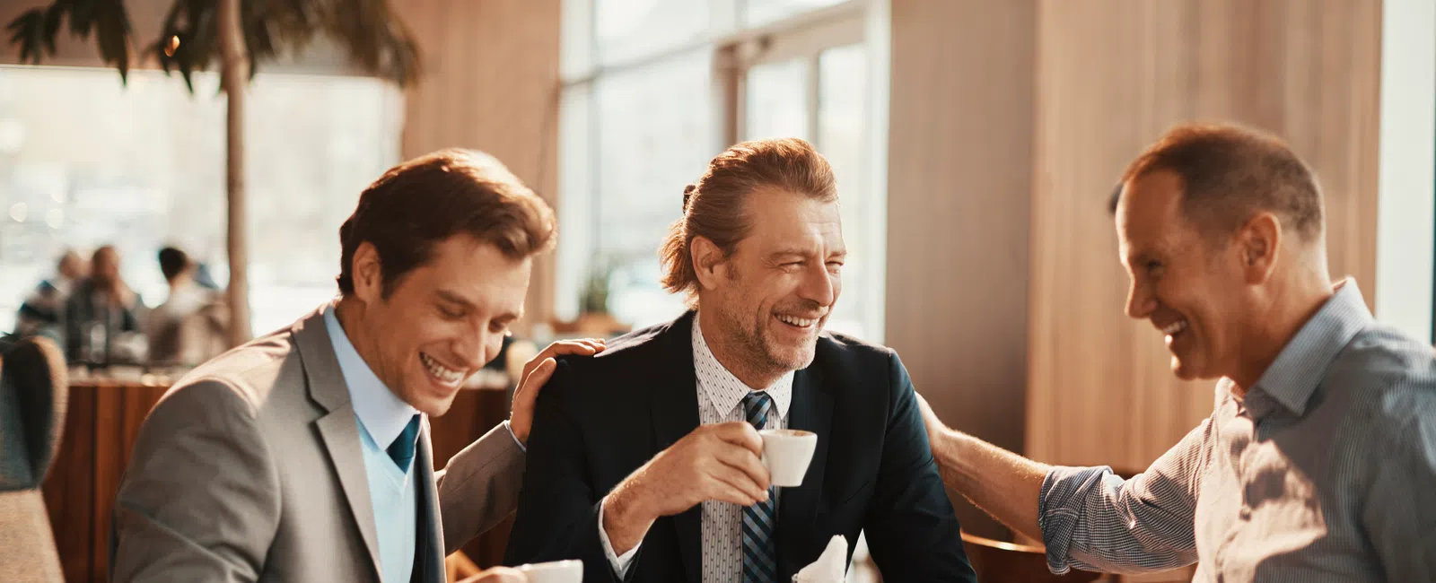 Three men in business attire enjoying coffee and smiling together in a bright, modern cafe setting