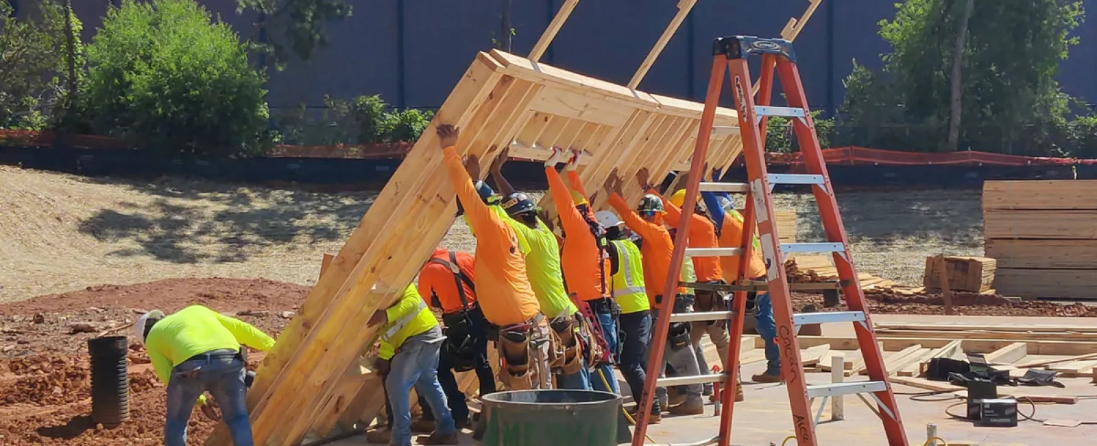 Construction workers in safety gear lifting a large wooden frame on a sunny job site with tools and ladder nearby