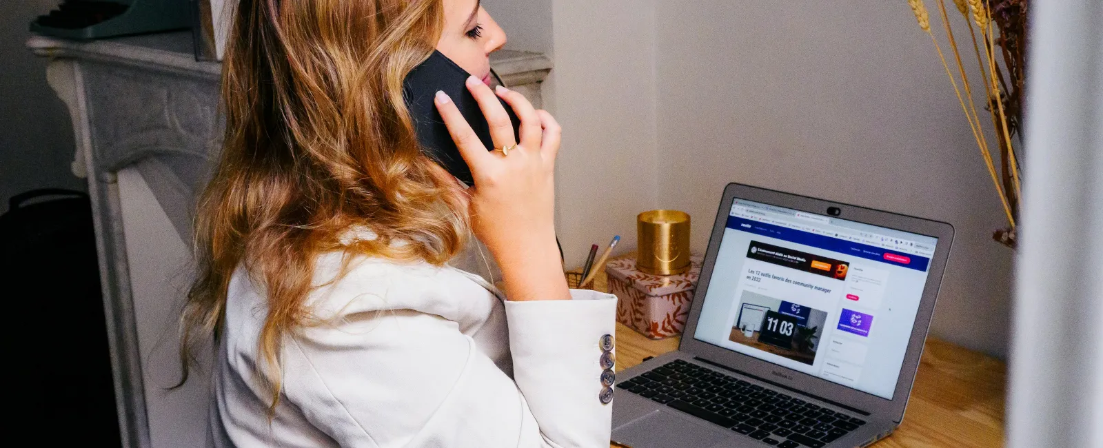 Woman in white blazer talking on a business phone while working on laptop at wooden desk indoors
