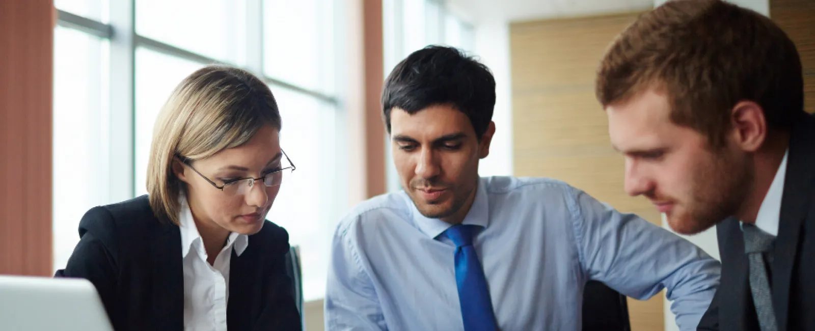 Three business professionals in formal attire collaborating around a tablet in a modern office.