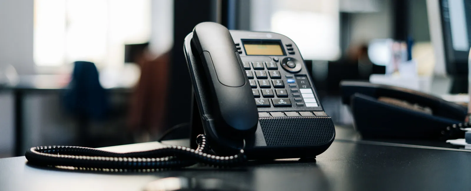 Black office desk phone with coiled cord on a desk in a modern workspace with blurred background.