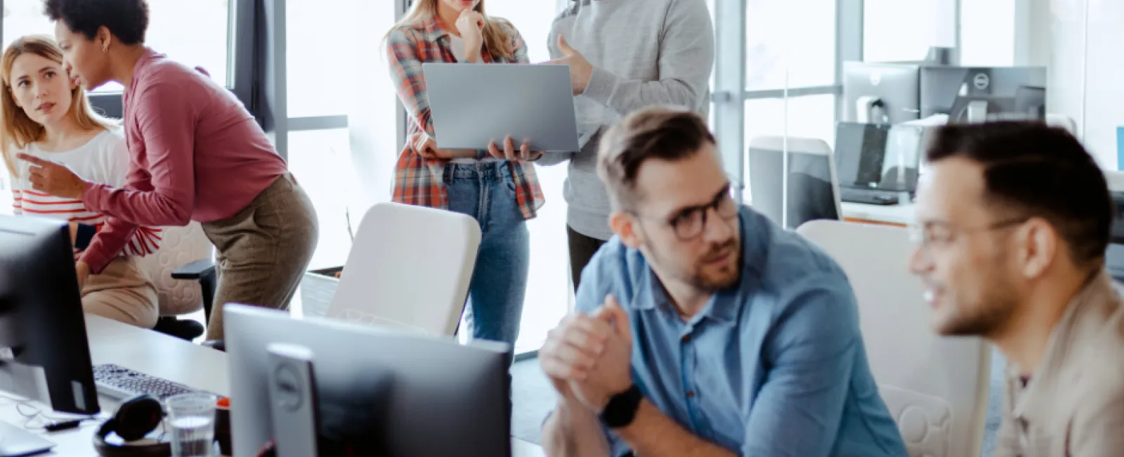 Diverse office team collaborating with laptops and computers in a bright modern workspace with large windows.