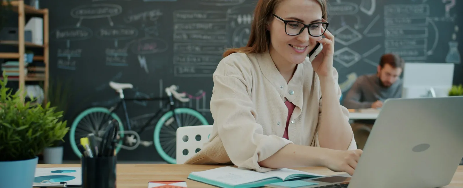 Young woman with glasses using laptop and talking on phone in creative workspace with chalkboard and bicycle.