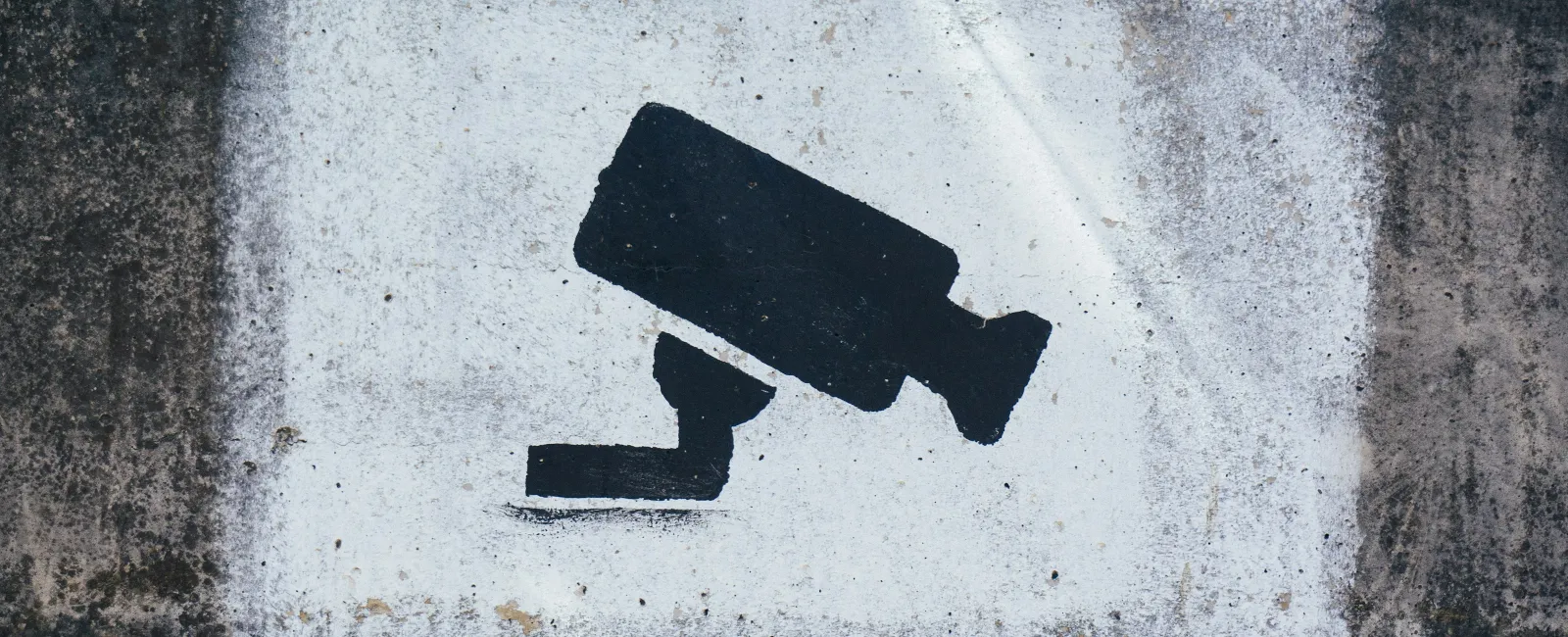 Black surveillance camera symbol painted on a weathered concrete wall with white background patch.