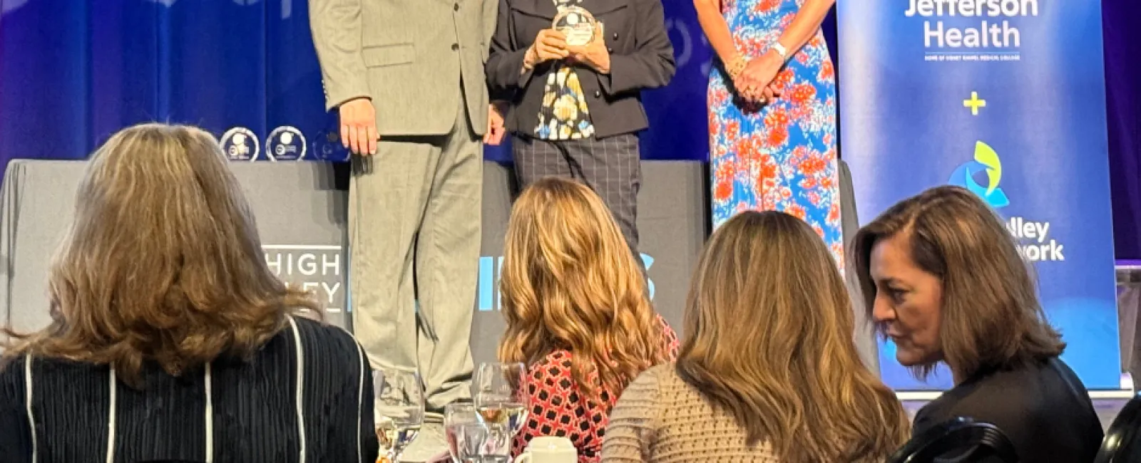 Three people standing on stage at a business women awards event with Jefferson Health banner and audience in foreground