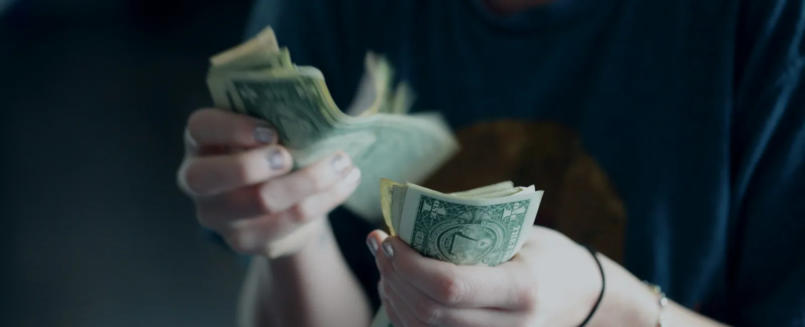 Person counting one-dollar bills with both hands wearing a dark shirt and a ring on the finger.