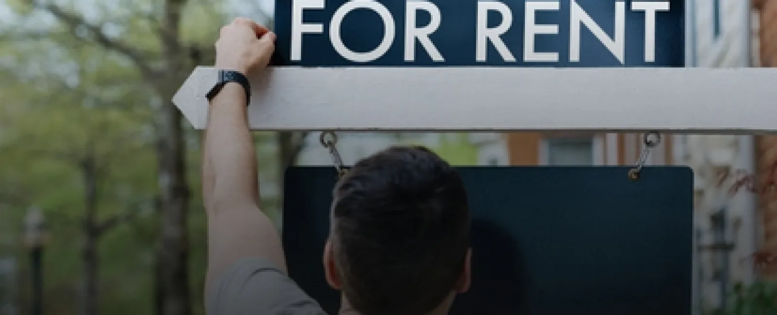 Person hanging a 'For Rent' sign outside a residential building on a tree-lined street.