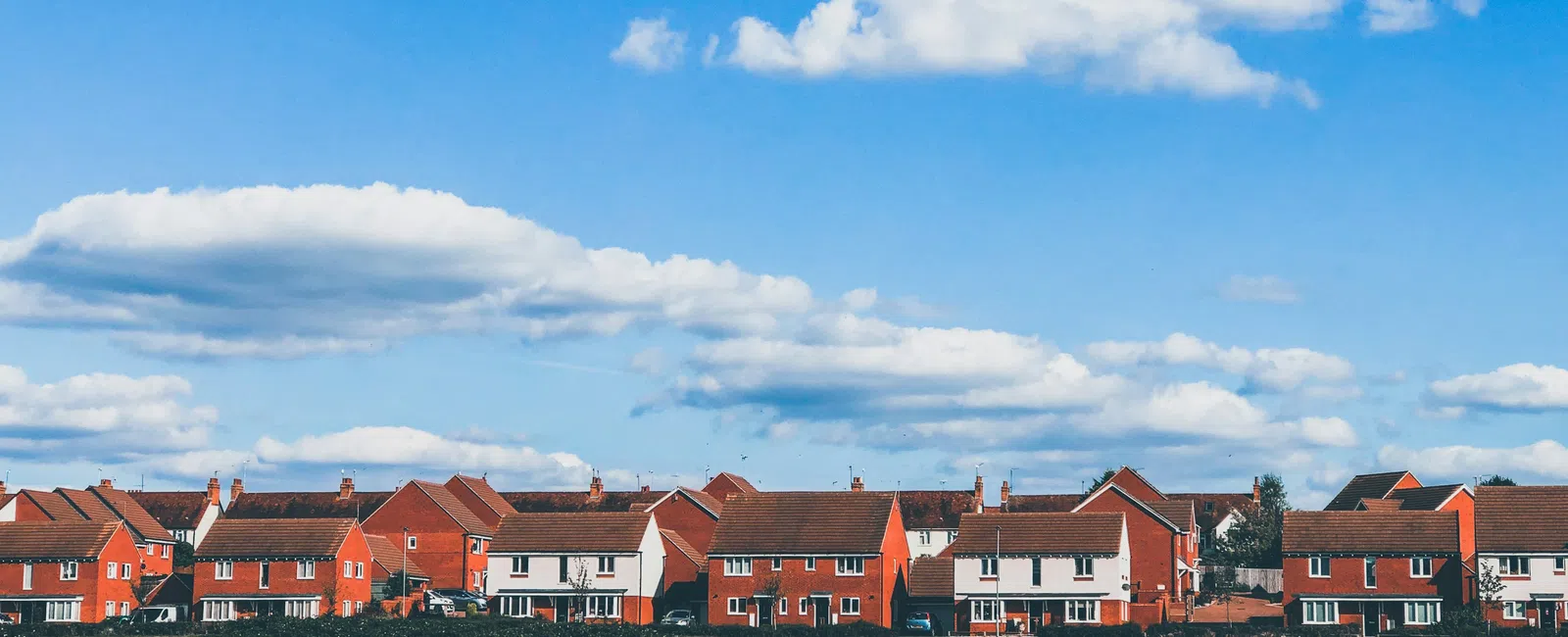 Row of suburban red brick houses under a bright blue sky with scattered white clouds and a green field in front.