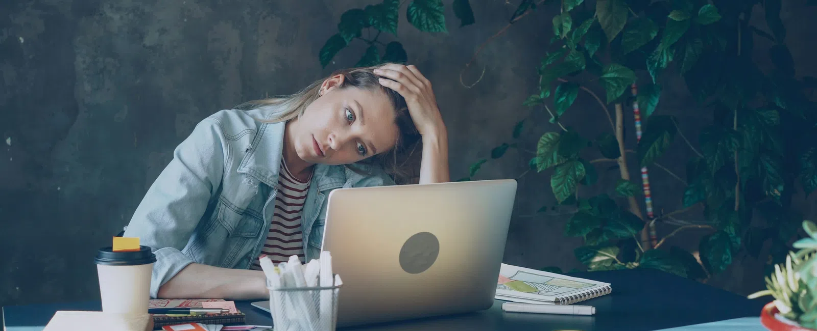 Tired woman working on laptop at desk with coffee and notebooks in indoor office with plants.