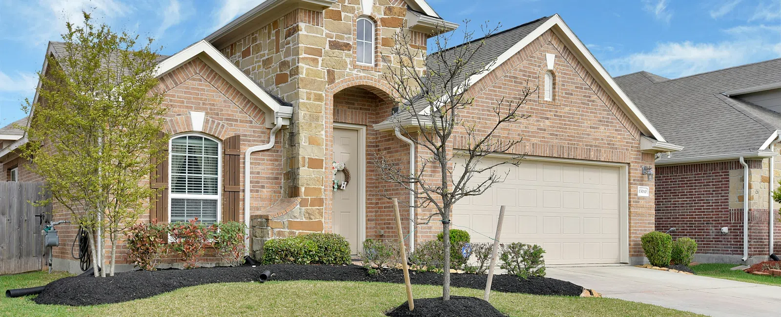 Brick and stone suburban house with a two-car garage and landscaped front yard under a blue sky.