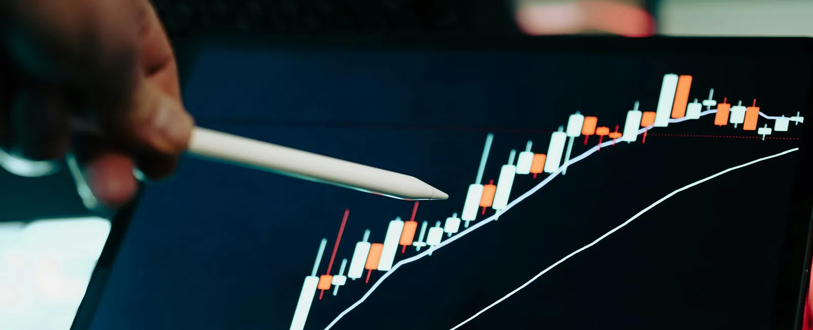 Person using a stylus to analyze an upward trending candlestick chart on a tablet screen for stock trading.