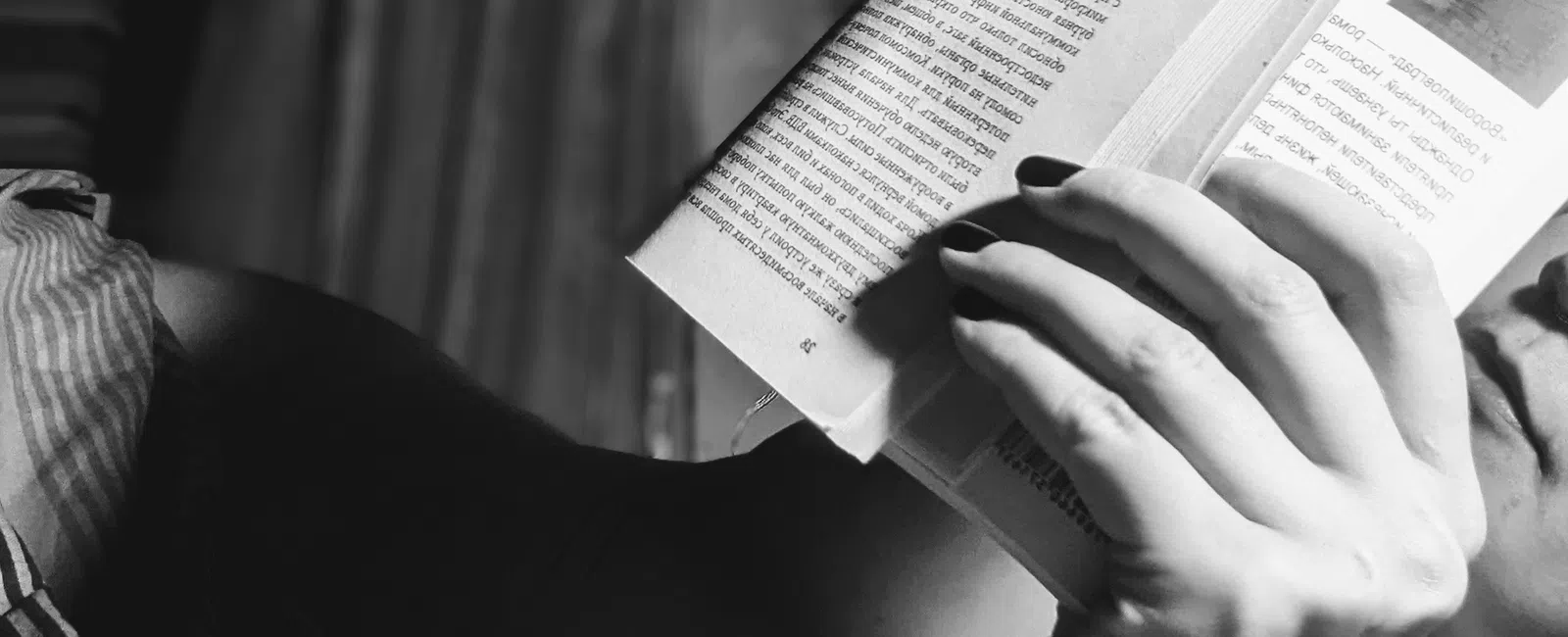 Black and white photo of a person reading a book closely, focusing on hand and part of face in soft light.