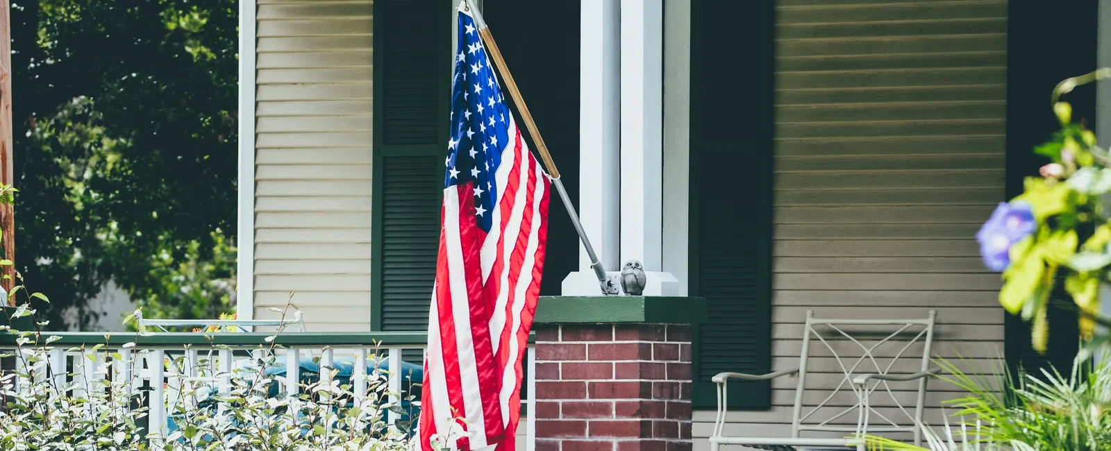 American flag displayed on a front porch with brick pillar, green plants, and outdoor chair in daylight.