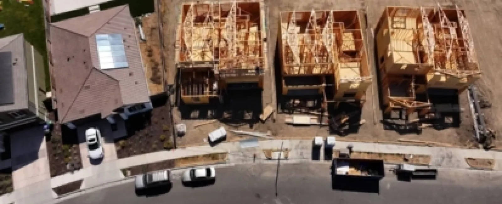 Aerial view of a residential street showing three houses under construction with wooden frames beside completed homes.