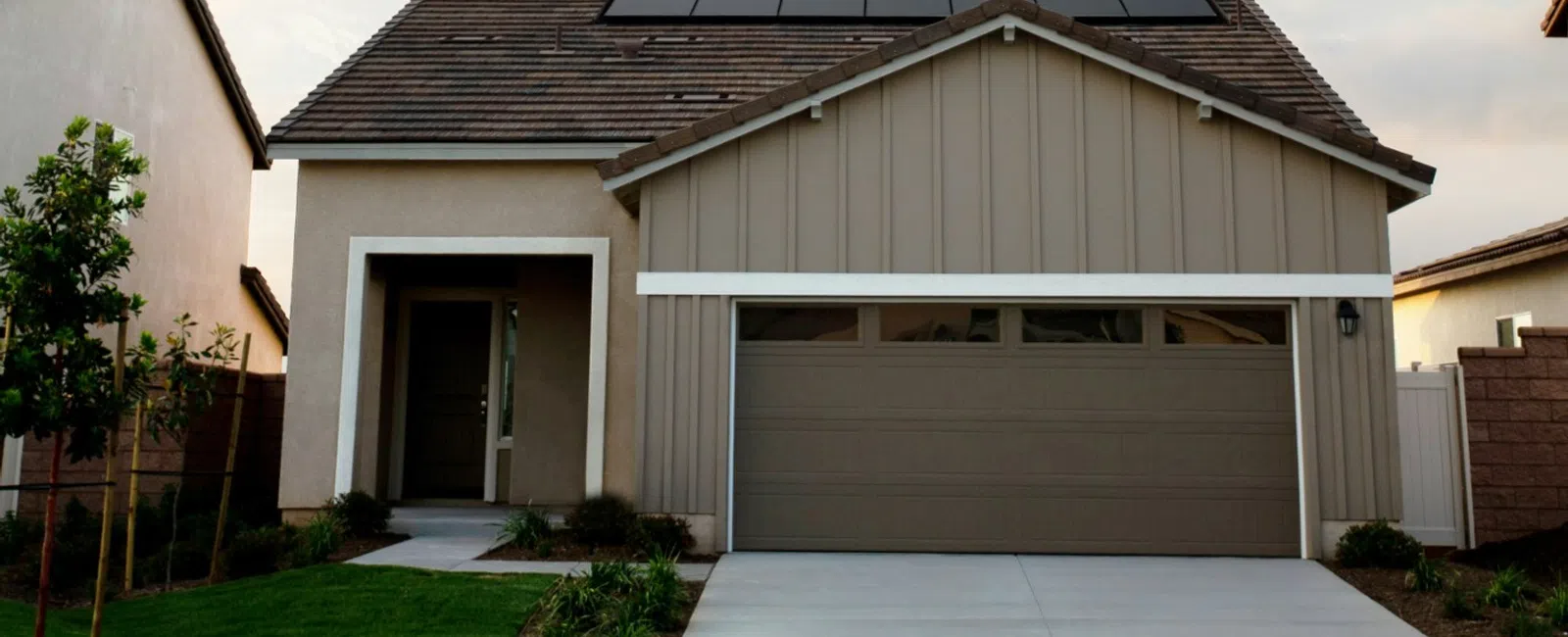 Modern suburban house with solar panels on roof, beige exterior, driveway, and landscaped front yard under clear sky.