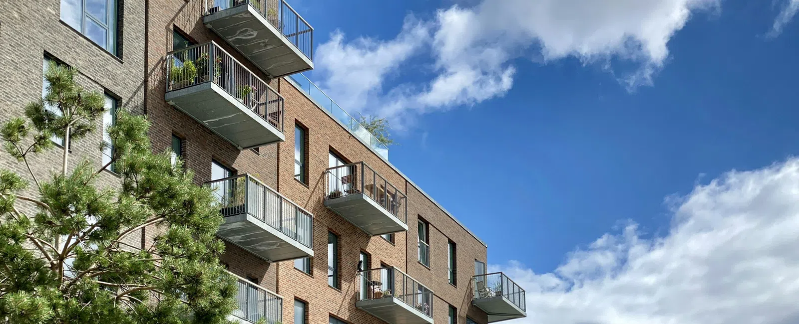 Modern brick apartment building with balconies under a blue sky with scattered clouds and a tree in the foreground