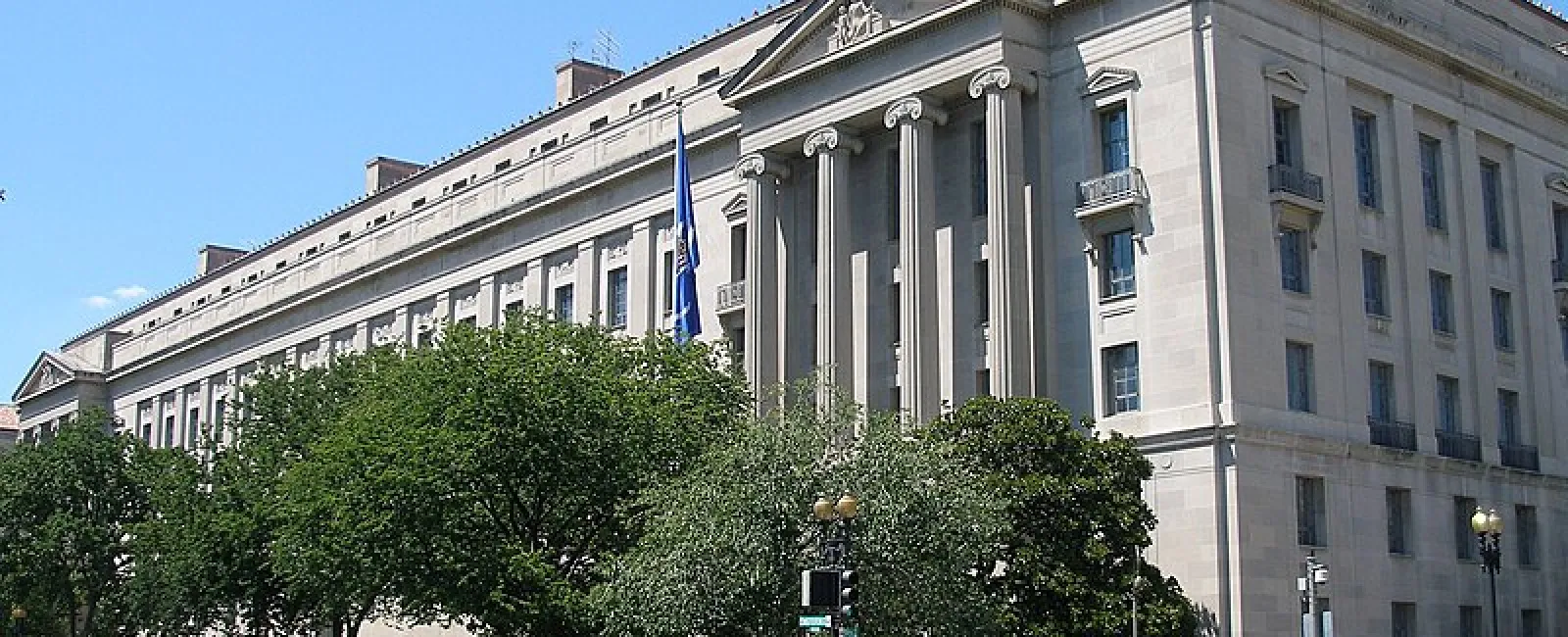 Historic government building with tall columns and lush trees lining the street under a clear blue sky