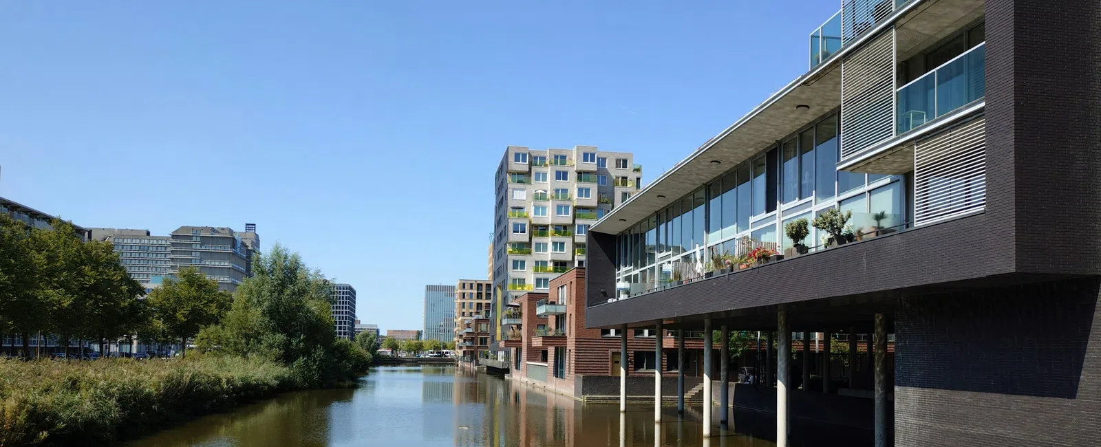 Modern buildings reflect in a river under clear blue sky with greenery on the opposite bank of the waterway.