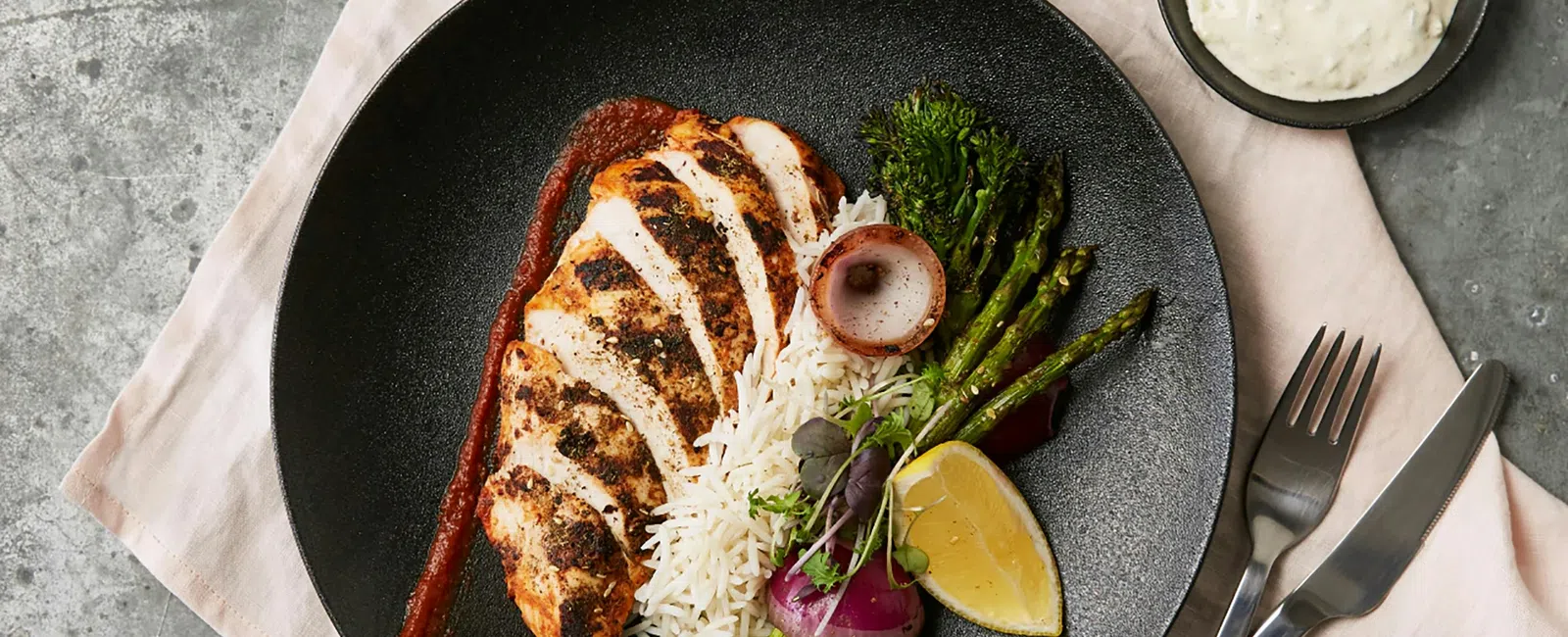 Grilled chicken breast with rice, asparagus, lemon wedge, roasted onion, fresh herbs on black plate with sauce and cutlery