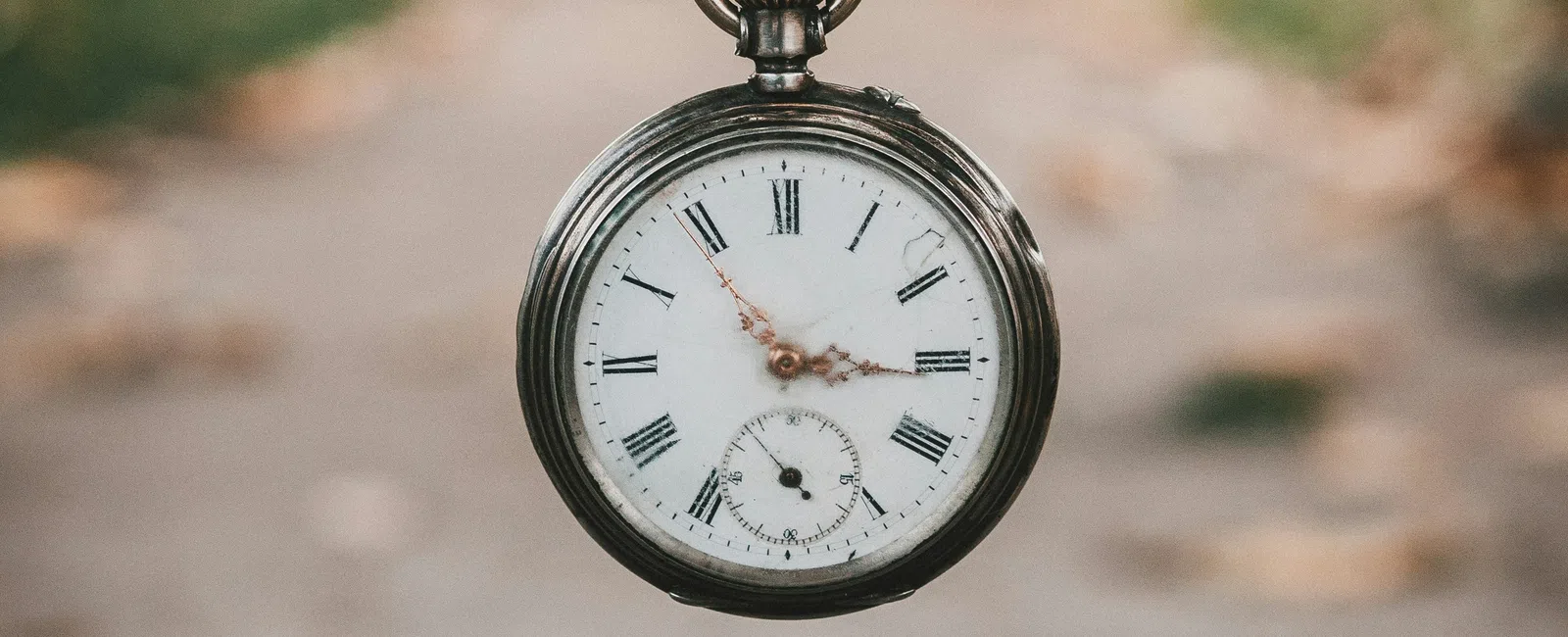 Vintage pocket watch with Roman numerals hanging on a chain outdoors with blurred autumn background.