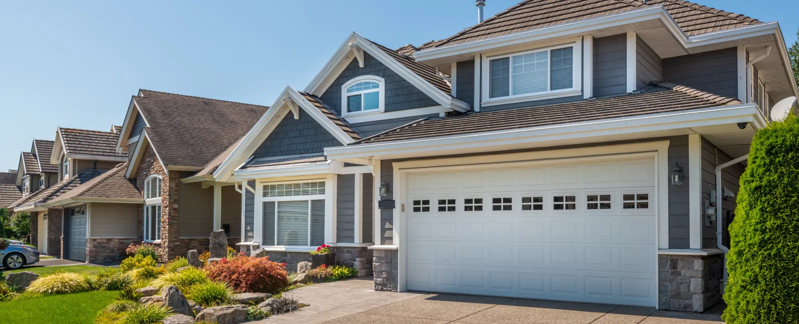 Modern suburban house with gray siding, white garage door, landscaped front yard, and clear blue sky.