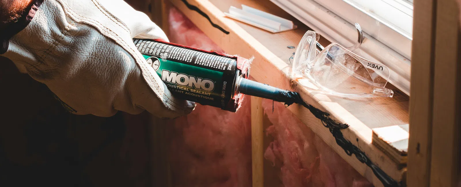 Worker applies black sealant around a wooden window frame with insulation and safety glasses nearby.
